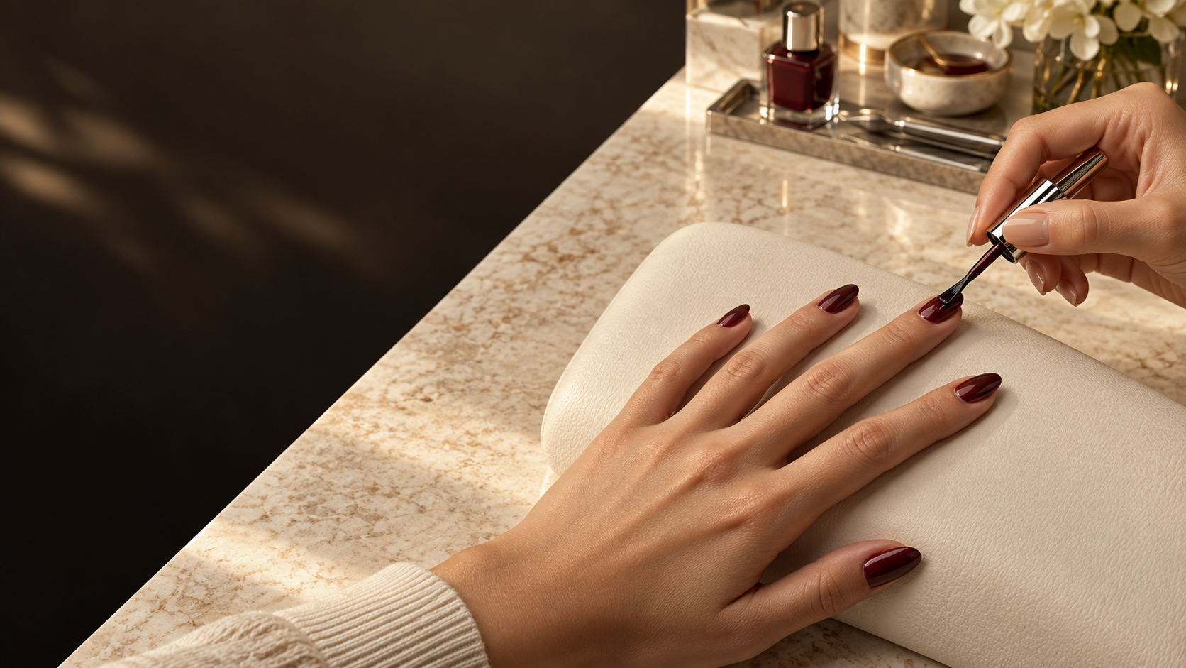 Una mano con cinco dedos mientras una manicurista aplica esmalte color vino.
